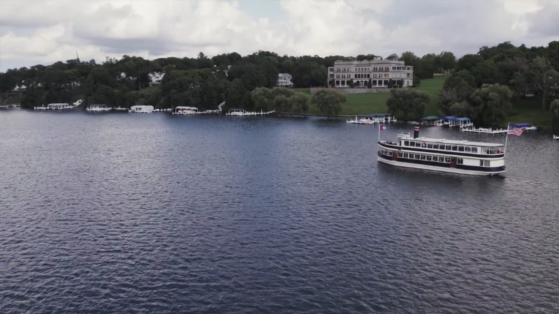 A Boat Is Cruising on Lake Geneva in Wisconsin, with Lakeside Homes and Greenery in The Background