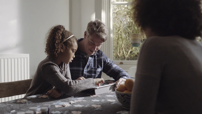 A father helps his daughter with homework at the kitchen table as the mother looks on