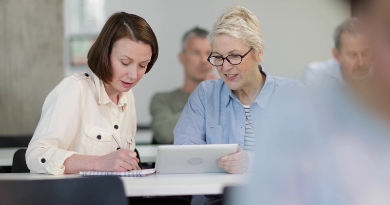 Two adult learners collaborate during professional development, one writing in a notebook while the other uses a tablet
