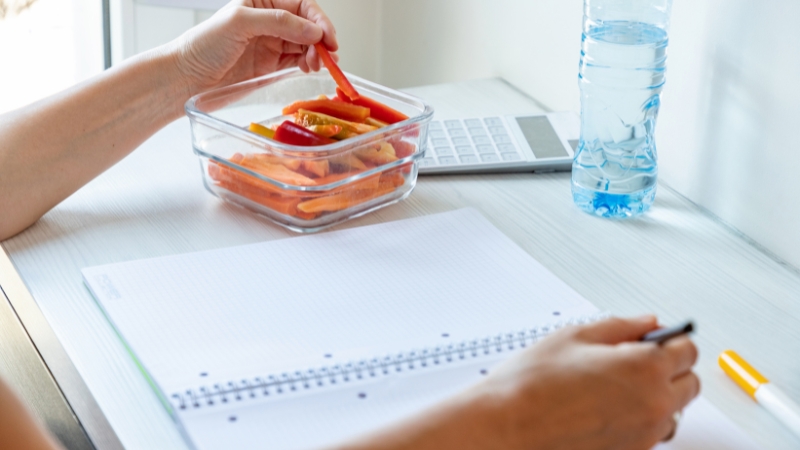 A person studying at a desk with a notebook, eating sliced vegetables from a glass container and a bottle of water nearby