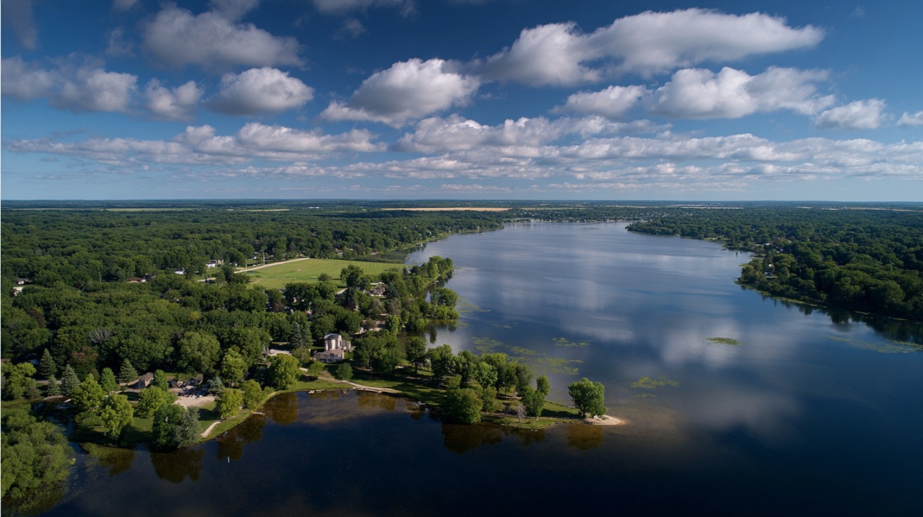 Aerial view of Shawano Lake in Wisconsin, showing calm blue water surrounded by green forests, lakeside homes, and a few sandy shores under a sky with scattered clouds