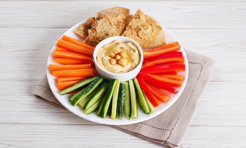A vibrant platter of fresh-cut carrots, cucumbers, and red bell peppers arranged around a bowl of creamy hummus
