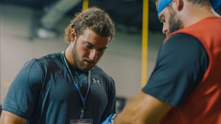An athletic trainer helps an athlete inside a gym or training facility, with one person wearing a lanyard and dark gray shirt, and another in a red vest and blue headband