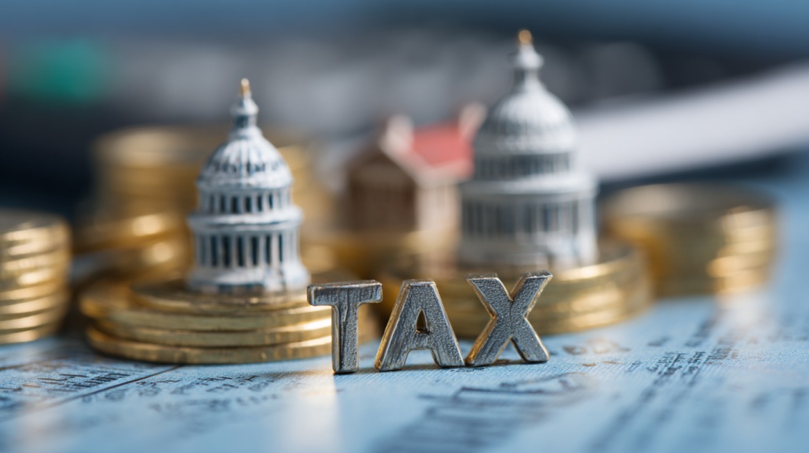 Stacks of coins with miniature government buildings and metal letters spelling the word "TAX" on a document background