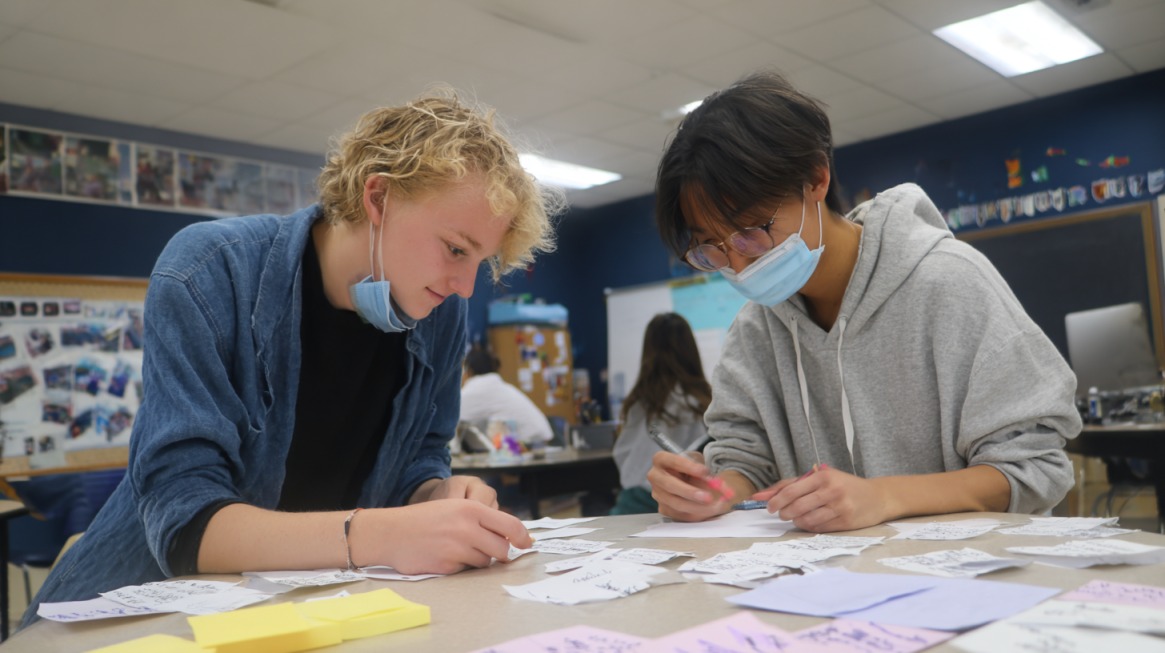 Two students working together at a classroom table with papers and notes