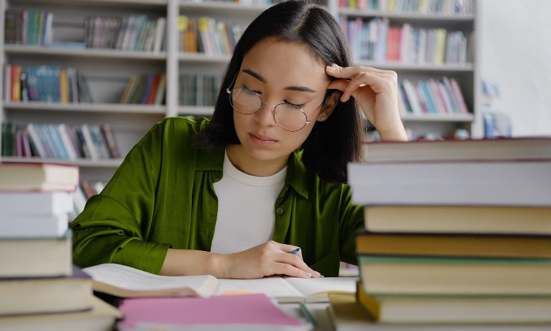 A woman with glasses sits at a desk surrounded by books