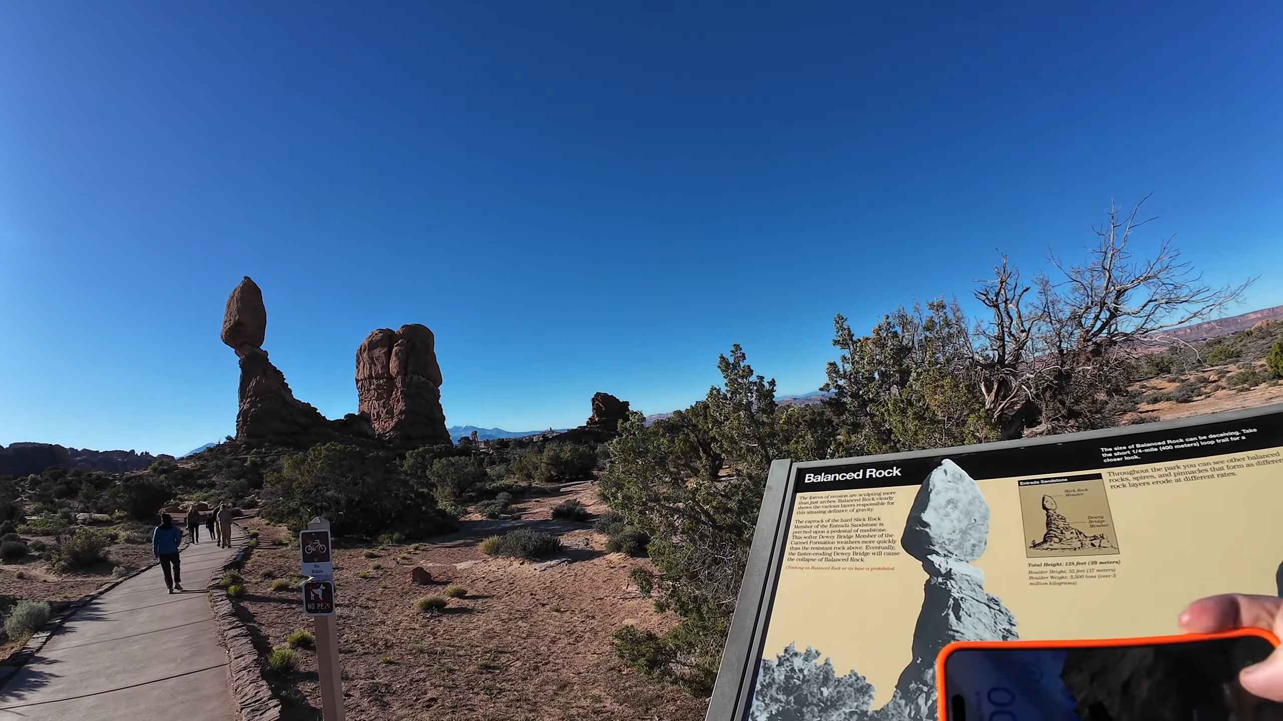 Visitors walking along a trail toward a tall balanced rock under a clear blue sky