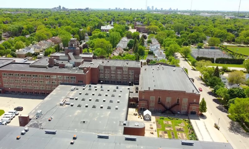 Aerial view of a large brick school building with a flat roof surrounded by green trees and residential houses