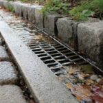 Close-up of a street curb with cobblestone pavement and a metal storm drain grate carrying rainwater