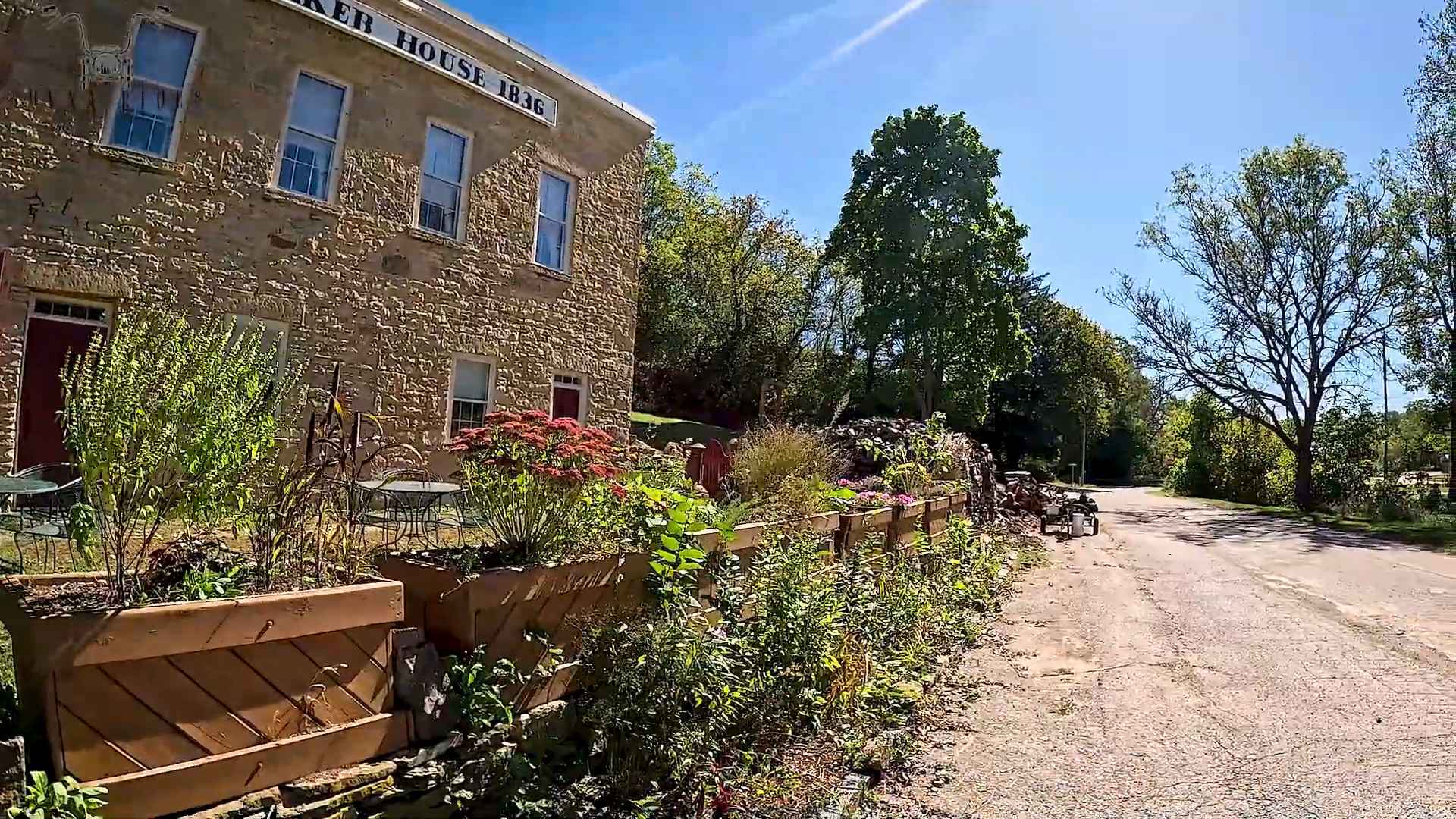 Stone building labeled “Hotel House 1836” beside a quiet country road with flower boxes and trees in bloom