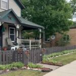 Cozy house with a porch and picket fence beside a small-town street with a U.S. post office in the background