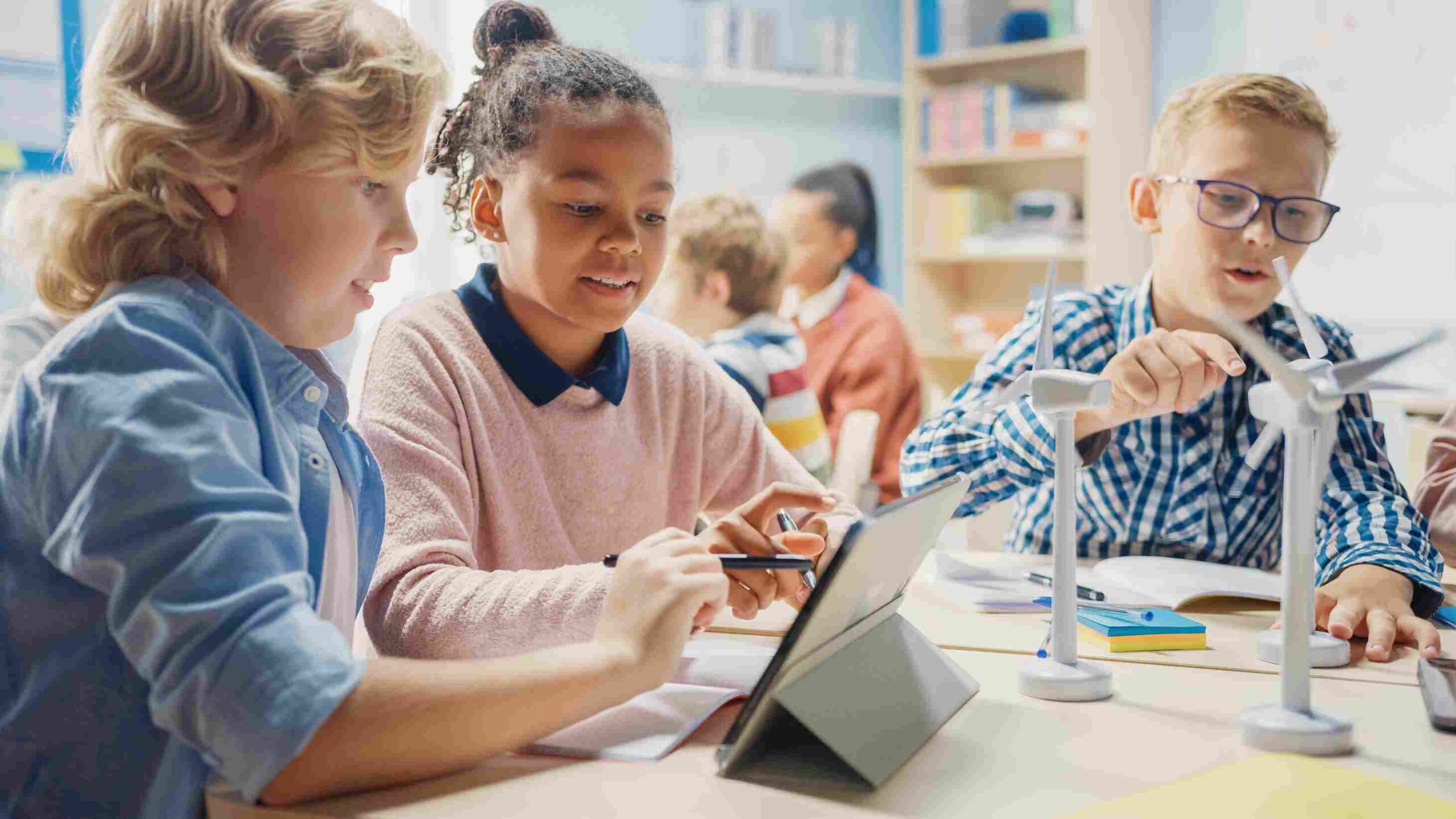 Students working together on a tablet with small wind turbine models on a classroom desk