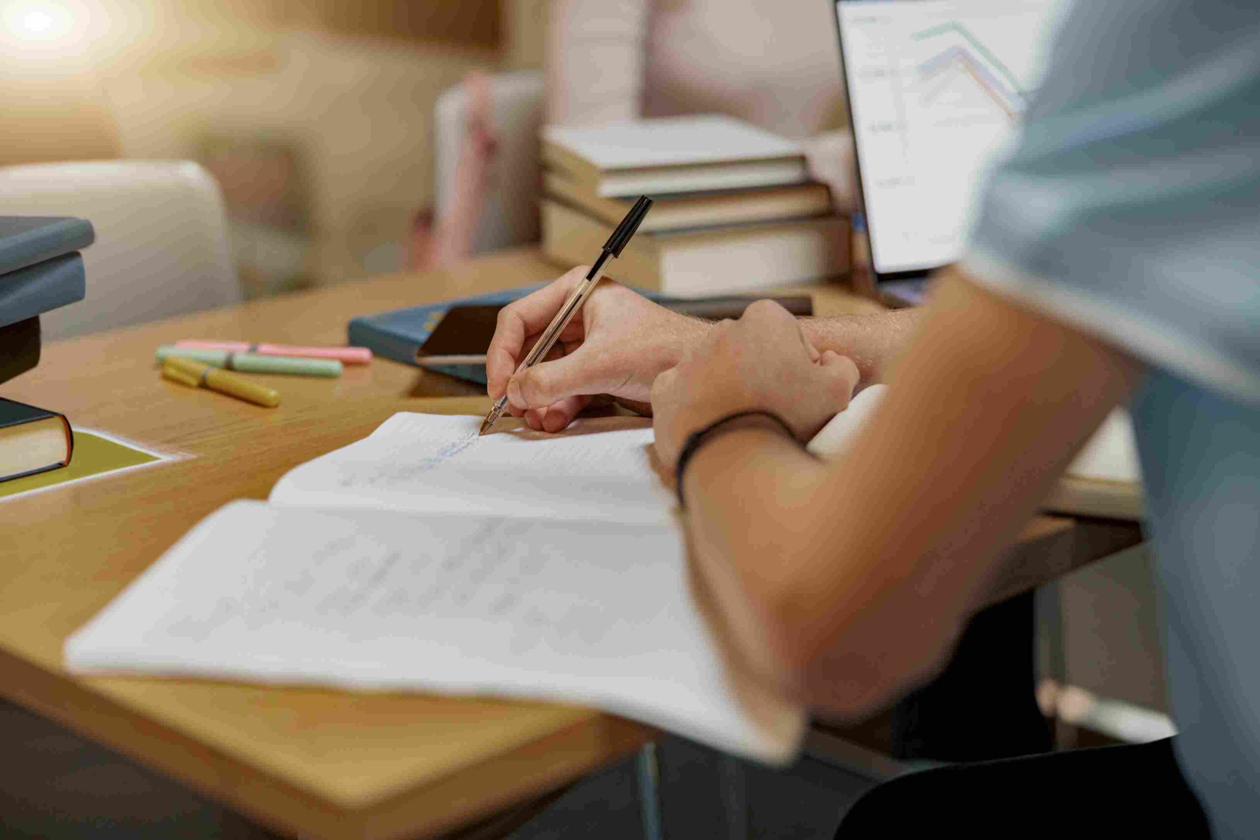 Student writing notes at a desk with books and a laptop nearby