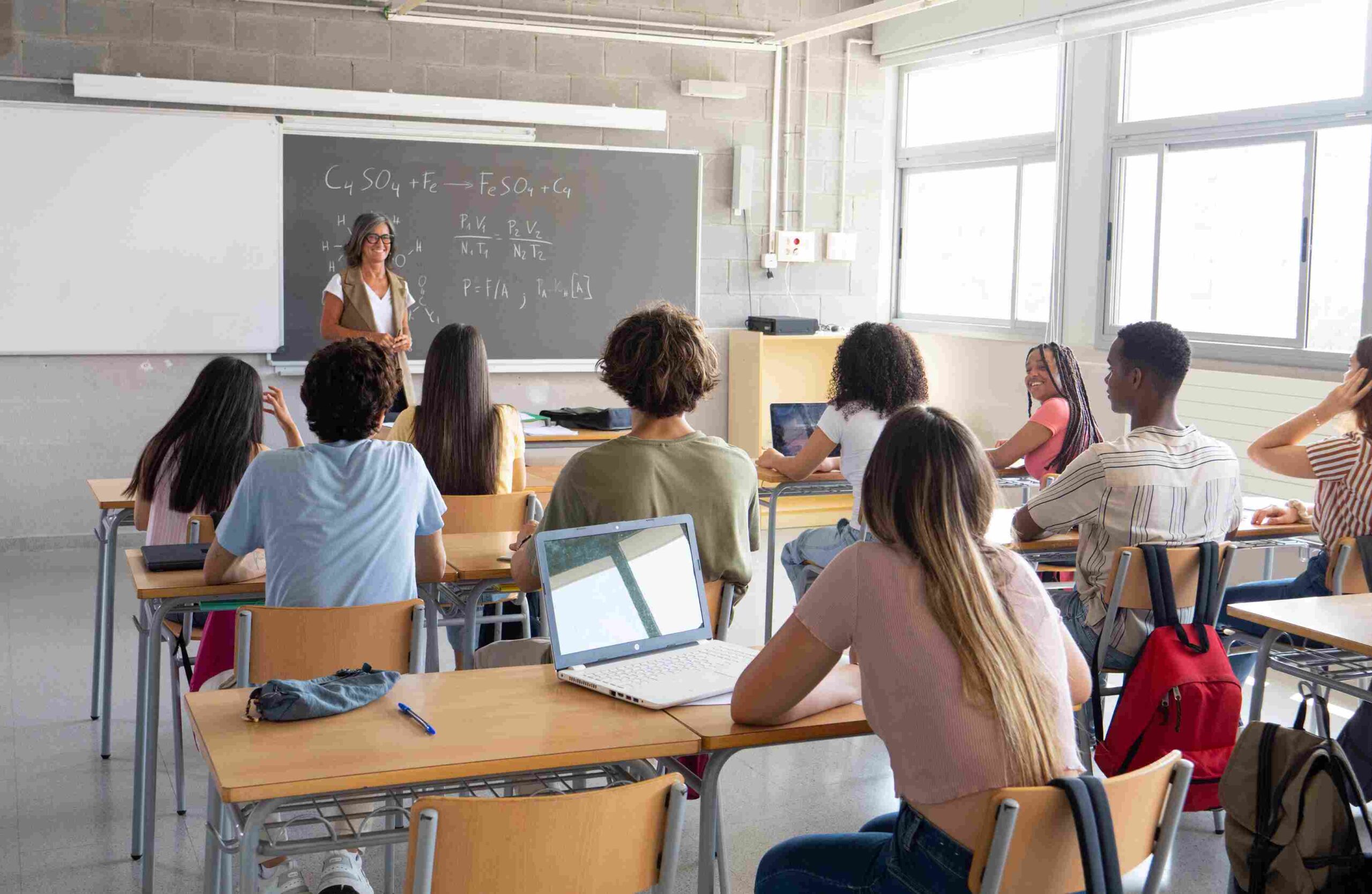 Teacher leading a lesson in a classroom with students seated at desks and natural light from large windows
