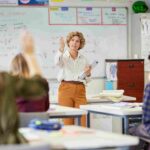 Teacher standing at the front of a classroom leading a lesson with students raising hands